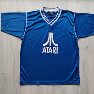 Könnte beinhalten: Ein kurzärmeliges blaues Trikot mit einem weißen Atari-Logo und Schriftzug auf der Brust. Der V-Ausschnitt ist mit einer weißen Borte verziert, die ein wiederkehrendes Muster aus kleinen blauen Dreiecken aufweist. Das Shirt hat weiße Paspeln an den Schultern.