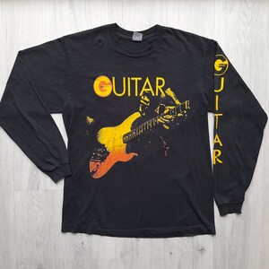 May include: Black long-sleeved t-shirt with a yellow and orange graphic of a person playing a guitar. The text "GUITAR" is printed in yellow on the front of the shirt. The word "GUITAR" is printed vertically on the left sleeve in yellow.