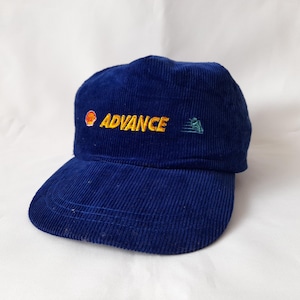 Könnte beinhalten: Marineblaue Cord-Baseballkappe mit dem gelb gestickten Wort "ADVANCE". Die Kappe hat auch ein Shell-Logo und eine kleine grüne Grafik. Der Hut hat einen gebogenen Schirm und eine strukturierte Krone.