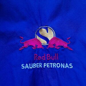 Puede incluir: Tela azul real con un logotipo de Red Bull bordado. El logotipo presenta dos toros rosas, un c&iacute;rculo dorado con la letra "S" y las palabras "Red Bull" y "SAUBER PETRONAS" bordadas en blanco.