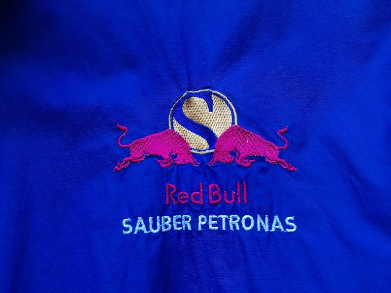 Puede incluir: Tela azul real con un logotipo de Red Bull bordado. El logotipo presenta dos toros rosas, un c&iacute;rculo dorado con la letra "S" y las palabras "Red Bull" y "SAUBER PETRONAS" bordadas en blanco.