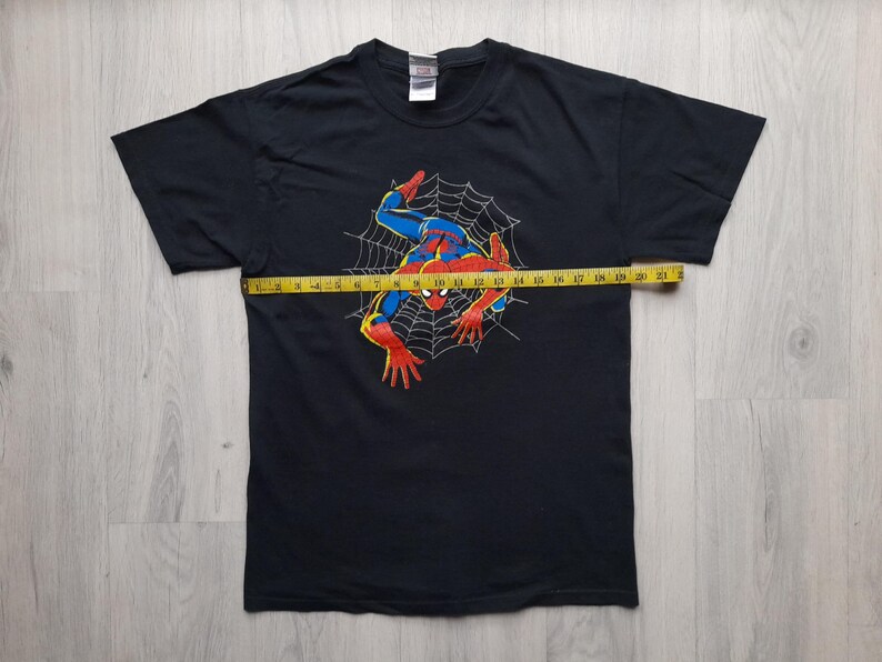 Pu&ograve; includere: T-shirt nero con grafica di Spider-Man in rosso, blu e giallo, su una ragnatela. Un metro a nastro giallo &egrave; posto sulla maglietta. Il design della maglietta &egrave; a tema supereroi.