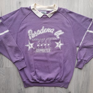 May include: Purple sweatshirt with white stripes on the sleeves and a white graphic print that says "Pasadena G. American System Supporter" with a star on each side of the text and a graphic of five people in a line.