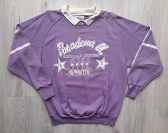 May include: Purple sweatshirt with white stripes on the sleeves and a white graphic print that says "Pasadena G. American System Supporter" with a star on each side of the text and a graphic of five people in a line.