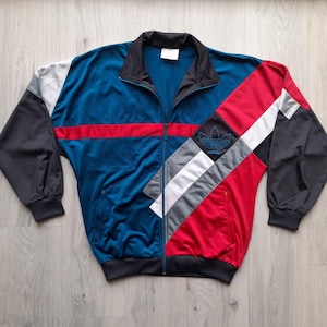 Peut inclure: Veste de survêtement vintage avec un design color block audacieux. Le corps est bleu sarcelle avec une bande rouge, accentué par des panneaux noirs, blancs et gris. Le logo Adidas est visible sur le panneau rouge. La veste a une fermeture éclair complète et un col noir.