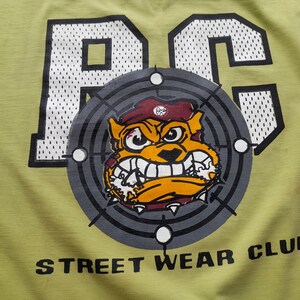May include: A green t-shirt featuring a white and black graphic of the letters "RC" with a cartoon bulldog wearing a red beret in a target design. The text "STREET WEAR CLUB" is printed below the graphic.