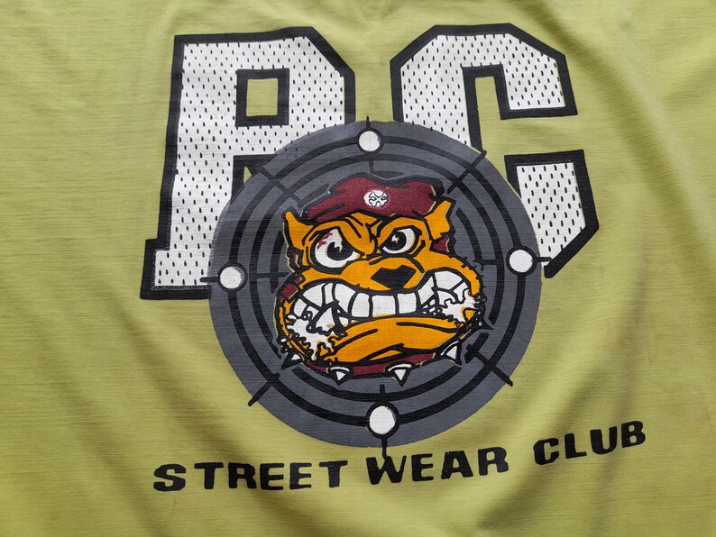 May include: A green t-shirt featuring a white and black graphic of the letters "RC" with a cartoon bulldog wearing a red beret in a target design. The text "STREET WEAR CLUB" is printed below the graphic.