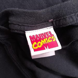 Pu&ograve; includere: Primo piano del colletto di una maglietta nera con un'etichetta Marvel Comics. L'etichetta &egrave; rettangolare con sfondo rosso e testo giallo. Il logo Marvel Comics &egrave; visibile.