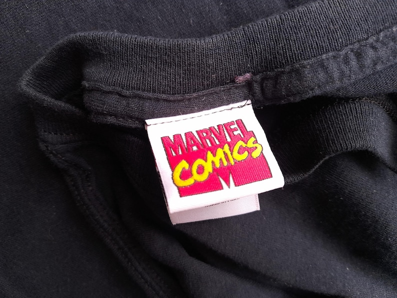 Pu&ograve; includere: Primo piano del colletto di una maglietta nera con un'etichetta Marvel Comics. L'etichetta &egrave; rettangolare con sfondo rosso e testo giallo. Il logo Marvel Comics &egrave; visibile.