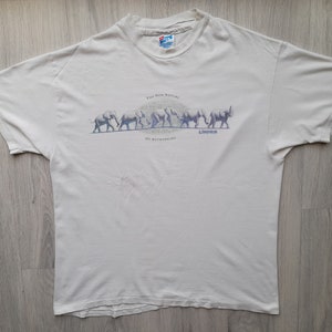 Puede incluir: Camiseta blanca con un gráfico de una línea de elefantes caminando. El texto "The New Species of Metamorphosis" está impreso debajo de los elefantes.