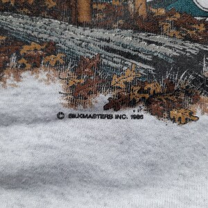May include: A close-up of a white t-shirt with a brown and green image of a cabin in the woods. The text "&copy; Silkmasters Inc. 1988" is printed on the bottom of the shirt.
