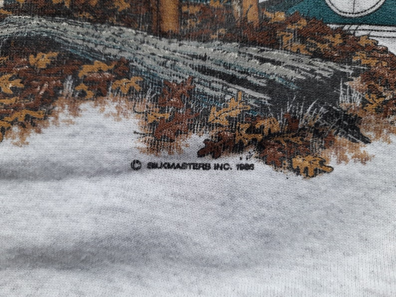 May include: A close-up of a white t-shirt with a brown and green image of a cabin in the woods. The text "&copy; Silkmasters Inc. 1988" is printed on the bottom of the shirt.