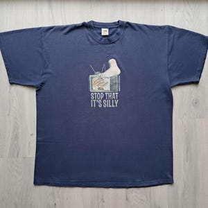 May include: A navy blue short-sleeve t-shirt featuring a graphic of a television set with a foot resting on top. The screen displays text and the words "STOP THAT IT'S SILLY" are printed in white below the television graphic on the front of the shirt.