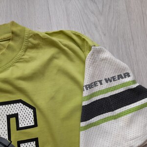 May include: A lime green t-shirt with a black and white mesh design on the sleeve. The sleeve has the text "Street Wear" printed on it. The front of the shirt has a large black and white mesh letter "C" on it.