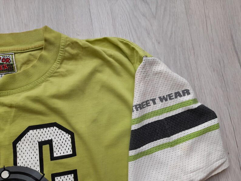 May include: A lime green t-shirt with a black and white mesh design on the sleeve. The sleeve has the text "Street Wear" printed on it. The front of the shirt has a large black and white mesh letter "C" on it.