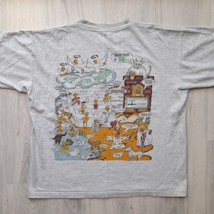 Puede incluir: Una camiseta gris con una ilustración de dibujos animados de una escalera al cielo. La ilustración incluye ángeles, un diablo y personas que suben las escaleras. El texto "Life After Death" está impreso en la escalera.
