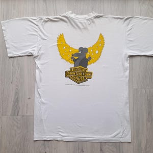 Puede incluir: Camiseta blanca con un diseño de ratón de dibujos animados. El ratón tiene alas de queso amarillo y sostiene queso. El texto de la camiseta dice "SULSTEIN'S BORN TO BE FUNNY MAUSE".