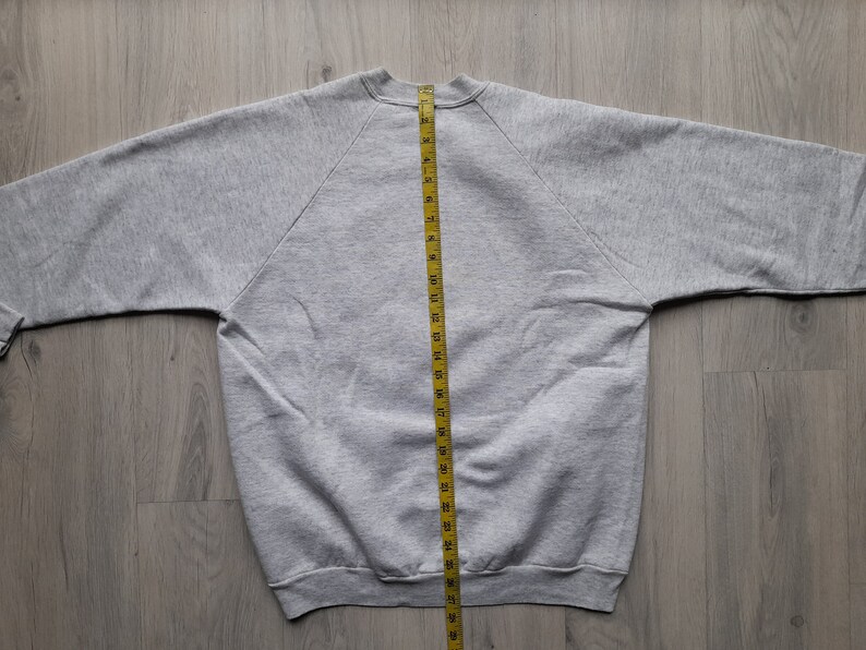 May include: A gray sweatshirt with a yellow measuring tape running down the center back. The sweatshirt is laid flat on a wooden surface.