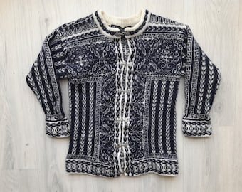 May include: A navy blue and white patterned cardigan sweater. The sweater features a round neckline, long sleeves, and a button-up front with decorative clasps. The intricate design includes geometric and floral patterns.