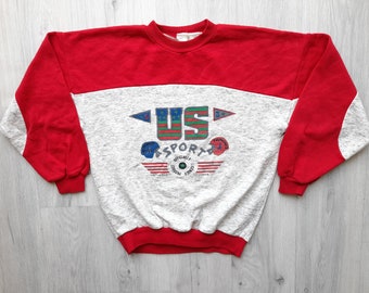 May include: Red and grey sweatshirt with a graphic print of "US SPORTA" with a flag design and stars. The sweatshirt has a crew neck and long sleeves.
