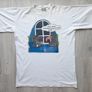 Puede incluir: Camiseta blanca con una ilustración de dibujos animados de dos ratones en la cama mirando por una ventana. La ventana tiene una parte superior arqueada y un fondo azul. El texto en un bocadillo dice: "LIEST KEINE GEBRAUCHSANWEISUNG DABEI ?"