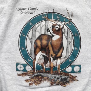 May include: A gray sweatshirt with a graphic print of a white-tailed deer in a forest setting. The deer has large antlers and is standing on a log. The graphic is surrounded by a teal blue circle with smaller deer silhouettes. The text "Brown County State Park" is printed above the graphic.