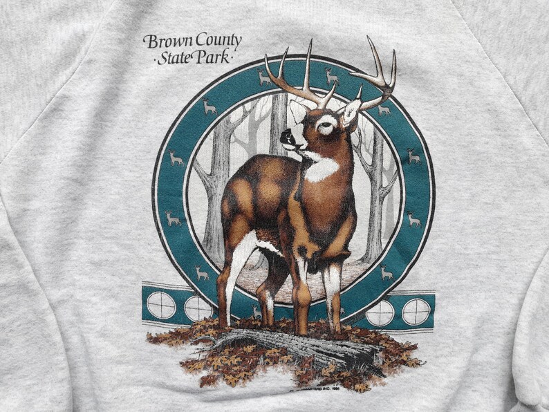 May include: A gray sweatshirt with a graphic print of a white-tailed deer in a forest setting. The deer has large antlers and is standing on a log. The graphic is surrounded by a teal blue circle with smaller deer silhouettes. The text "Brown County State Park" is printed above the graphic.