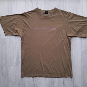 May include: A brown t-shirt with short sleeves and a crew neck. The text "NAKED YOUTH BEACHERS" is printed across the chest in a lighter shade of brown. The shirt is laid flat on a light wood-grain surface.