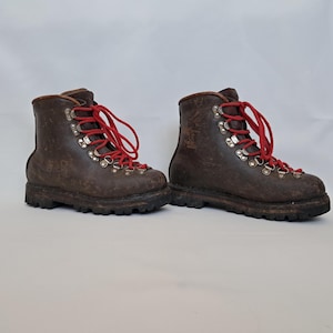 Op de afbeelding: Een paar donkerbruine leren wandelschoenen met felrode veters, zilverkleurige metalen oogjes en haken, en dikke, donkere rubberen profielzolen. De schoenen hebben een hoog model met een gewatteerde kraag en een textuur in het leer.