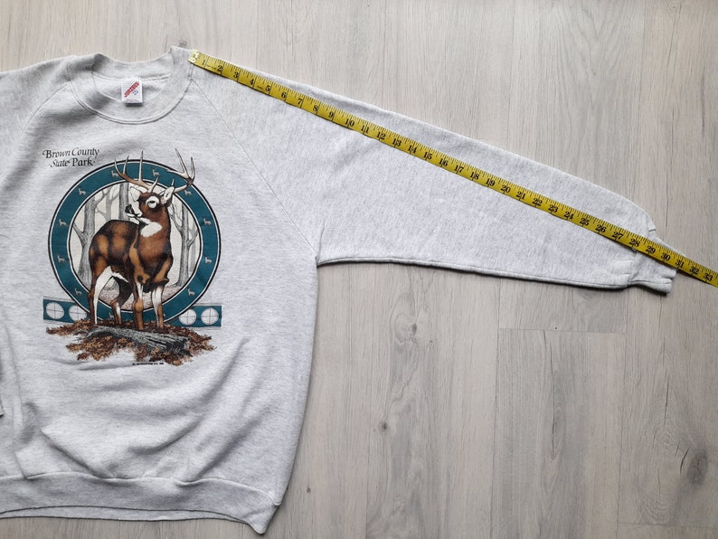 May include: Gray sweatshirt with a deer graphic and the text "Brown County State Park". The sweatshirt is laid flat with a measuring tape extending from the shoulder seam to the end of the sleeve, measuring 29 inches.