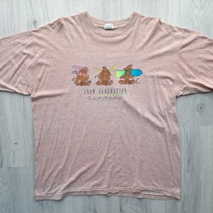 Peut inclure: Un t-shirt rose pâle avec un motif de trois singes portant des vêtements d'hiver. Le texte "SNOW GENERATION" est imprimé sous les singes. Le texte "Resort - Switzerland" est imprimé sous le texte "SNOW GENERATION".
