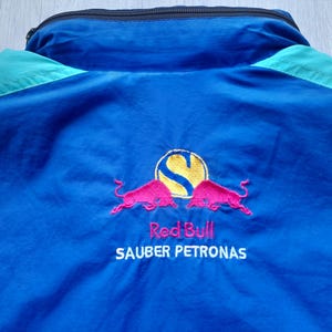 Puede incluir: Chaqueta azul y turquesa con cremallera negra y el logotipo de Red Bull Sauber Petronas bordado en la espalda. El logotipo presenta un c&iacute;rculo amarillo con una "S" estilizada y dos toros rosas.
