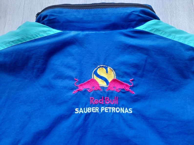 Puede incluir: Chaqueta azul y turquesa con cremallera negra y el logotipo de Red Bull Sauber Petronas bordado en la espalda. El logotipo presenta un c&iacute;rculo amarillo con una "S" estilizada y dos toros rosas.