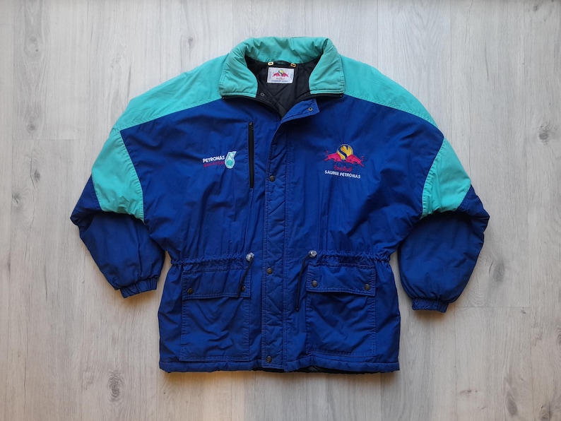Puede incluir: Chaqueta vintage azul, turquesa y negra con el logo de Petronas. La chaqueta tiene cuello y hombros turquesa, dos bolsillos delanteros y una cremallera negra. El texto "RACING PETRONAS" y un logotipo est&aacute;n en el pecho derecho.