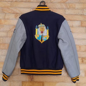 May include: Navy blue varsity jacket with gray sleeves and a yellow and black striped trim. The back features an embroidered crest with a lion wearing a crown. The jacket has a black collar with yellow stripes.