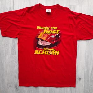 Pode incluir: Camiseta vermelha com um gráfico amarelo e preto de um piloto de corrida usando um capacete. O texto "Simply the best Simply SCHUMI" está impresso na camiseta.