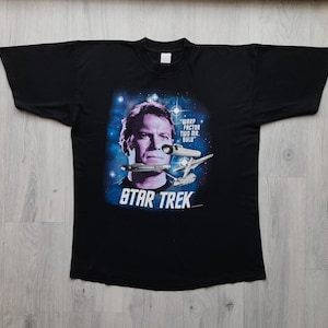 Puede incluir: Camiseta negra de Star Trek con un retrato del Capitán Kirk y el texto "Warp Factor Two Mr. Sulu".