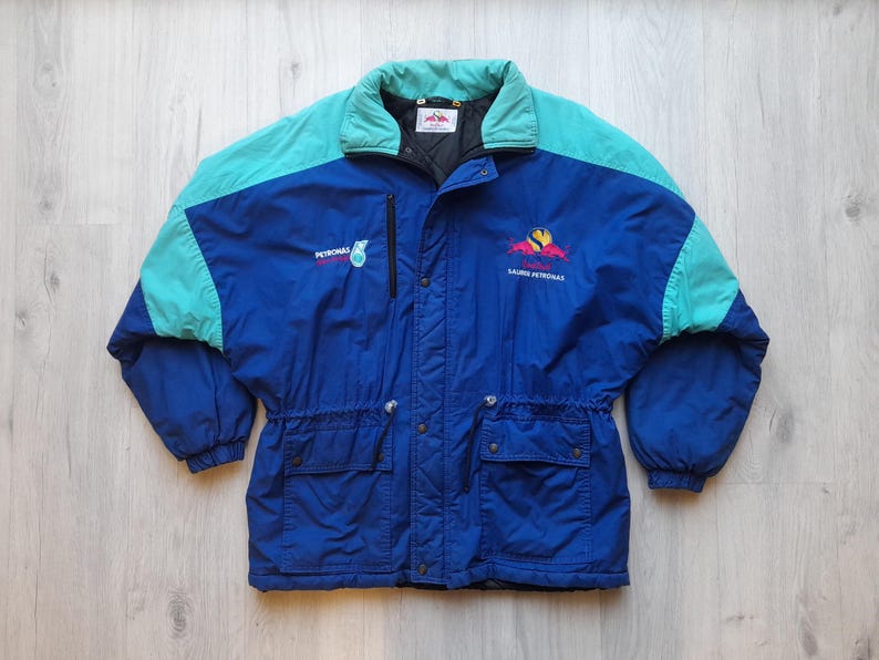 Puede incluir: Chaqueta vintage azul, turquesa y negra con los logotipos de Petronas y Saura Petronas. La chaqueta tiene cremallera, dos bolsillos delanteros y cuello y detalles en los hombros turquesa. Fabricada en material ligero.