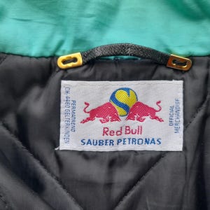 Puede incluir: Primer plano del interior de una chaqueta, con una etiqueta con el logotipo de Red Bull y el texto "SAUBER PETRONAS". La chaqueta tiene un forro acolchado negro, un cuello turquesa y un lazo de cuero negro con detalles dorados.
