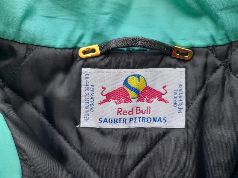 Puede incluir: Primer plano del interior de una chaqueta, con una etiqueta con el logotipo de Red Bull y el texto "SAUBER PETRONAS". La chaqueta tiene un forro acolchado negro, un cuello turquesa y un lazo de cuero negro con detalles dorados.