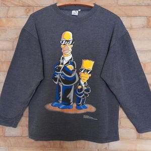Könnte beinhalten: Dunkelgraues Sweatshirt mit Homer und Bart Simpson mit Sonnenbrille und Anzügen. Die Zeichentrickfiguren stehen auf einer braunen Oberfläche. Das Sweatshirt hat lange Ärmel und einen Rundhalsausschnitt.