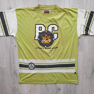 May include: A lime green t-shirt with a black and white graphic of a cartoon dog wearing a military cap. The graphic says "PC" and "Street Wear Club". The shirt has white and black stripes on the sleeves and bottom.