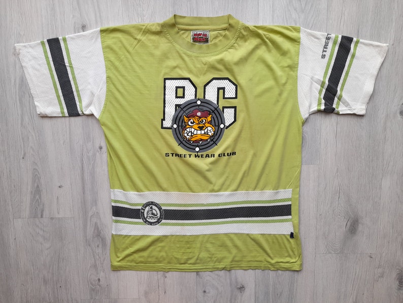 May include: A lime green t-shirt with a black and white graphic of a cartoon dog wearing a military cap. The graphic says "PC" and "Street Wear Club". The shirt has white and black stripes on the sleeves and bottom.