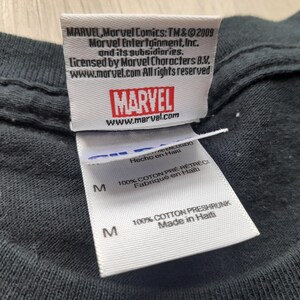 Pu&ograve; includere: Primo piano di un'etichetta di una maglietta nera con il logo Marvel e informazioni sul copyright. L'etichetta include il testo "100% Cotton Preshrunk Made in Haiti."