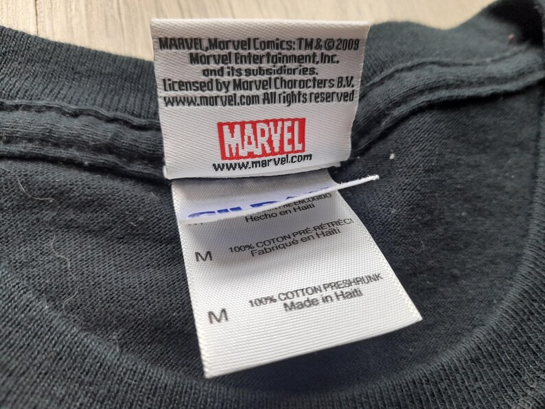 Pu&ograve; includere: Primo piano di un'etichetta di una maglietta nera con il logo Marvel e informazioni sul copyright. L'etichetta include il testo "100% Cotton Preshrunk Made in Haiti."