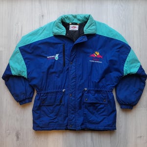 Puede incluir: Chaqueta vintage azul y turquesa con los textos "PETRONAS" y "LAURE PETRONAS". La chaqueta tiene cuello y hombros turquesa, dos bolsillos delanteros y cintura ajustable. Fabricada con material resistente al agua, es ideal para actividades al aire libre.