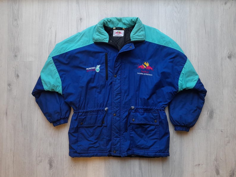 Puede incluir: Chaqueta vintage azul y turquesa con los textos "PETRONAS" y "LAURE PETRONAS". La chaqueta tiene cuello y hombros turquesa, dos bolsillos delanteros y cintura ajustable. Fabricada con material resistente al agua, es ideal para actividades al aire libre.