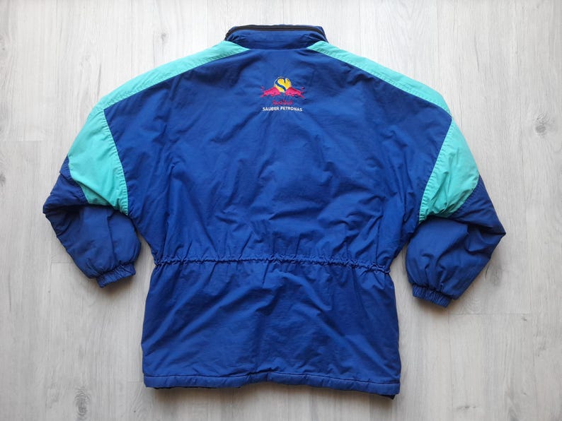 Puede incluir: Chaqueta vintage azul y turquesa con cintura con cord&oacute;n y cuello alto. La chaqueta tiene detalles en los hombros en color turquesa y un logotipo en la espalda. La chaqueta est&aacute; hecha de un material ligero.