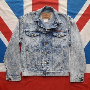 May include: A light blue denim jacket with a faded acid wash effect. The jacket has a button closure, two chest pockets, and two front pockets. The Wrangler logo is visible on the left chest pocket.