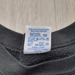 May include: A blue care label with white text detailing the fabric composition and care instructions for a garment. The label reads: "50% Cotton/Coton 50% Polyester Machine Wash Warm Colors Wash Separately Chlorine Bleach When Needed Do Not Iron Or Dry Clean Emblem Russell Corp Alexander City AL 35015 WPL 7232"